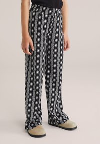 Pantalone širokih nogavica crno-bijelog uzorka s valovima i motivima sunca, izrađeni od laganog materijala. U kombinaciji s bež cipelama.