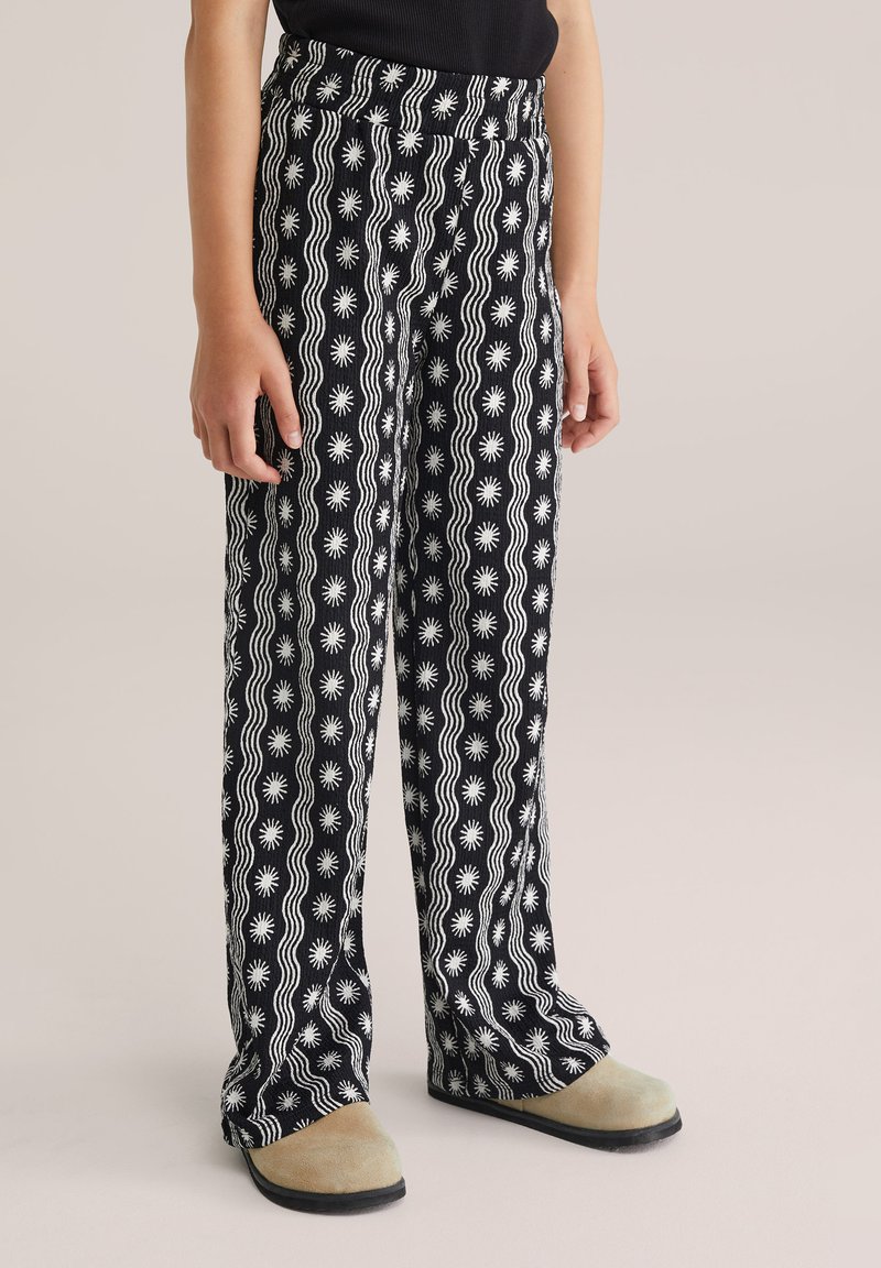 Pantalone širokih nogavica crno-bijelog uzorka s valovima i motivima sunca, izrađeni od laganog materijala. U kombinaciji s bež cipelama.