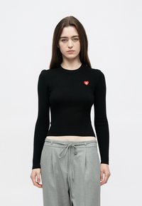 Top preto de manga comprida com nervuras, decote redondo e um pequeno coração vermelho no lado esquerdo do peito, combinado com calças cinzentas largas.