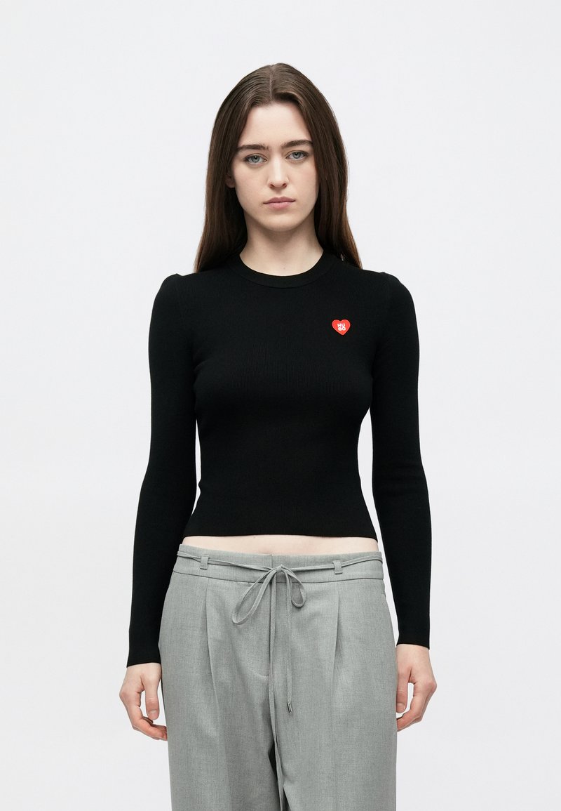 Top preto de manga comprida com nervuras, decote redondo e um pequeno coração vermelho no lado esquerdo do peito, combinado com calças cinzentas largas.