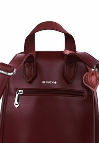 Burgundy faux leather backpack with two top handles, a zippered front pocket, and a heart-shaped charm. Stamped brand logo above pocket.
