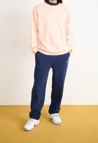 Ljusrosa sweatshirt med rund hals och logotypdetalj, kombinerad med marinblå joggers. Vita sneakers med ljusblå accenter fullbordar outfiten.