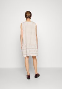 Ärmelloses beigefarbenes Kleid mit einem strukturierten Muster und einem Volantsaum, das horizontale Streifen und einen lässigen Schnitt aufweist. Getragen mit dunklen Schuhen.