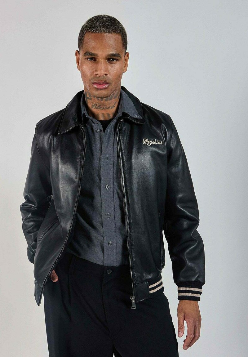 Veste bomber en cuir noir avec un col, des poignets côtelés rayés et un détail brodé "Redshine". Portée par-dessus une chemise grise à boutons.