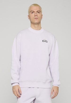 Giovane con capelli biondi corti indossa felpa e pantaloni bianchi, con piccolo logo nero "UNFAIR" sul petto a sinistra.