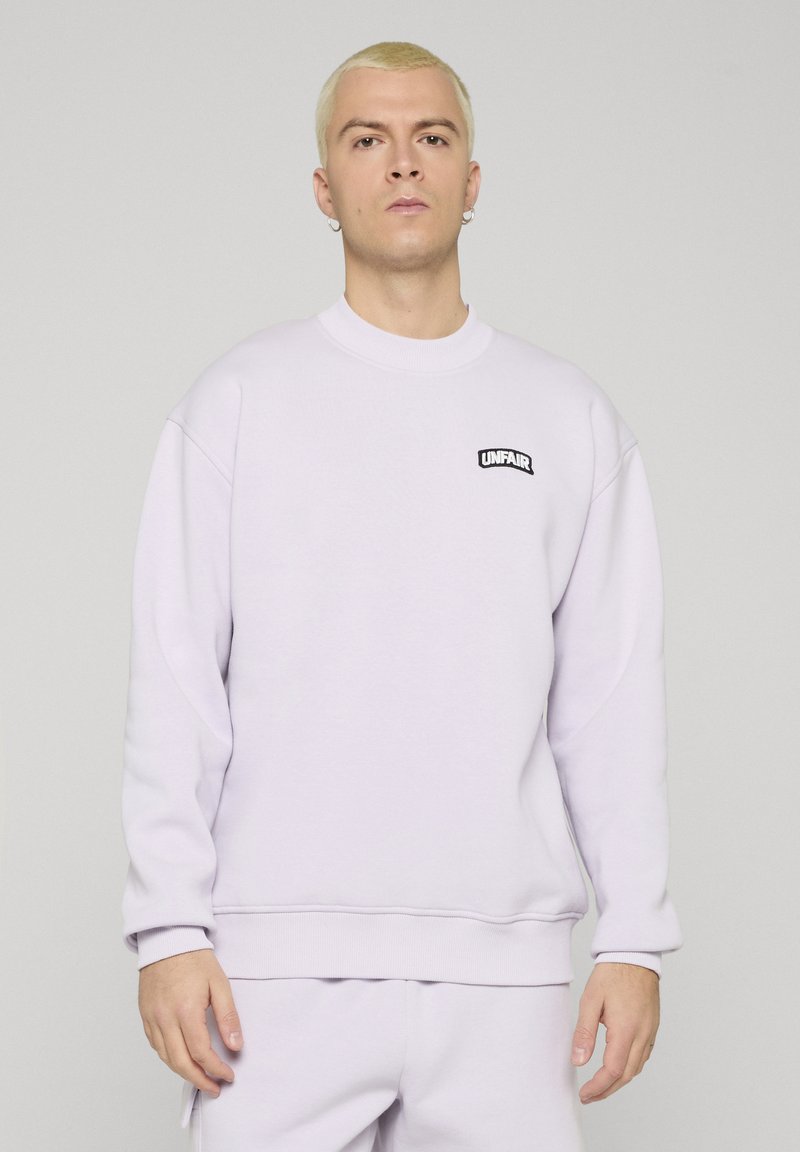 Noor inimene lühikeste blondide juustega kannab valget dressipluusi ja pükse, dressipluusi vasakul rinnal on väike must "UNFAIR" logo.