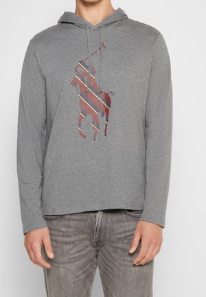 Hombre con sudadera con capucha gris con un logo de jugador de polo a rayas en rojo y azul, combinado con jeans grises desgastados sobre un fondo liso.