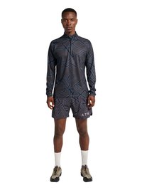 Mężczyzna w czarnej koszuli z długim rękawem na zamek oraz dopasowanych szortach z geometrycznym wzorem, białe skarpetki i beżowe buty do wędrówek, stojący.