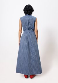 Langes Jeanskleid mit senkrechten Streifen, ärmellosem Design, tailliertem Gürtel und hohem Kragen. Tragen mit roten Schuhen. Rückansicht.