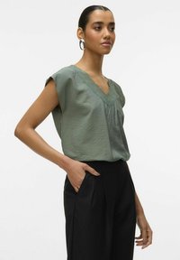 Blusa verde oliva con scollo a V e rifiniture in pizzo, dotata di maniche corte e vestibilità ampia, abbinata a pantaloni neri su misura.