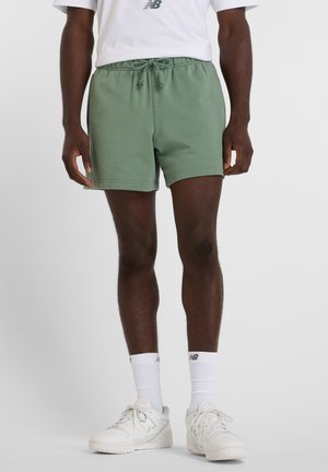 Mann trägt olivgrüne Shorts mit Kordelzug, weiße Sportsocken und weiße New Balance Sneaker, steht vor einfachem Hintergrund.