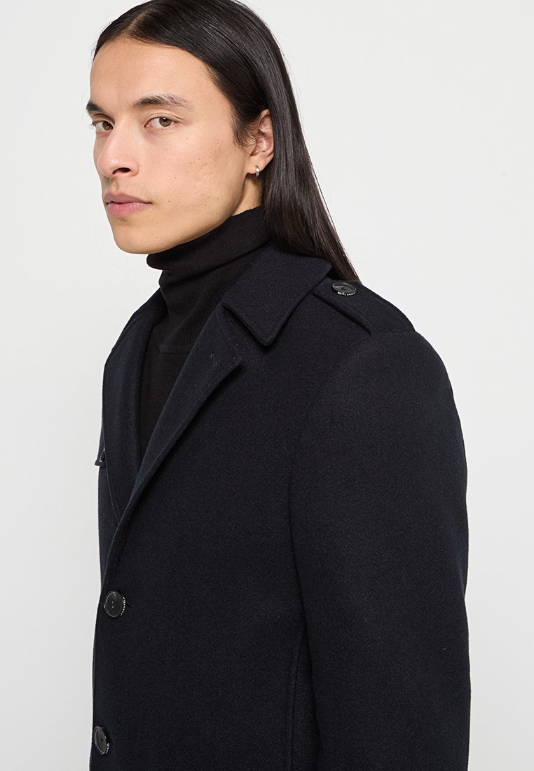 Cappotto di lana navy con ampio colletto, chiusure a bottone e tasche laterali. Texture liscia con vestibilità su misura, caratterizzato da un bottone decorativo sulla spalla.