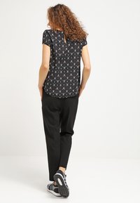 Mujer con cabello rizado vestida con una blusa negra de manga corta con estampado, pantalones negros y zapatillas en blanco y negro, de pie frente a un fondo blanco.