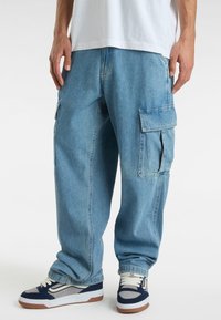 Pantaloni cargo in denim blu chiaro con molteplici tasche laterali, vestibilità ampia e polsini elasticizzati, abbinati a sneaker blu navy e grigie.