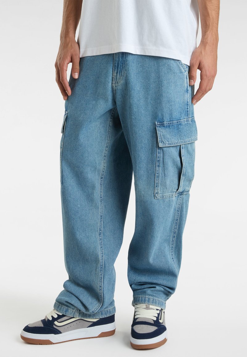 Pantaloni cargo in denim blu chiaro con molteplici tasche laterali, vestibilità ampia e polsini elasticizzati, abbinati a sneaker blu navy e grigie.