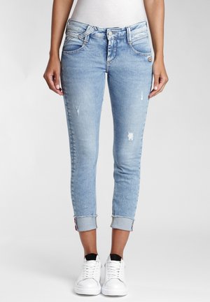 Femme portant un jean skinny bleu clair retroussé avec des détails légèrement usés et des baskets blanches, debout devant un fond neutre.