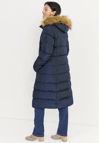 Marineblauwe donzen jas met een capuchon afgezet met bont, met een verticale doorgestikte structuur en elastische manchetten, gecombineerd met een spijkerbroek en beige laarzen met hak.