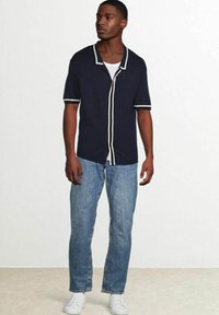 Marineblauwe korte mouwen overhemdd met crèmekleurige accenten, gecombineerd met lichtblauwe jeans en witte sneakers, tegen een neutrale achtergrond.