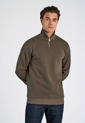 Herre zippered pullover i olivengrønn, laget av mykt stoff. Har en høy krage og ribbet fald, kombinert med mørke denimjeans.