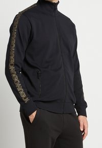 Chaqueta negra con cremallera hecha de tela suave, que presenta un cuello alto, un bolsillo lateral con cremallera y un logo dorado a lo largo de la manga. Puños acentuados.