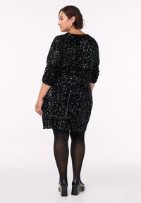 Sort kjole med palietter, lange ærmer, en tætsiddende talje og en kort kant. Bæres med sorte polka dot strømpebukser og sorte blokkede hæle.