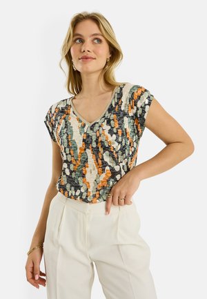 Mujer con una blusa de manga corta y escote en V con un patrón abstracto multicolor, combinada con pantalones blancos de talle alto, posando con una mano en el bolsillo.