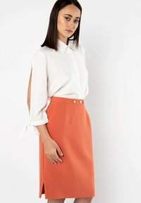 Witte textuur blouse met opgerolde mouwen, gecombineerd met een knielange oranje rok met vijf decoratieve knopen en zijopeningen.