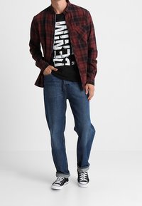 Rotes und schwarzes kariertes Langarmhemd über einem schwarzen T-Shirt mit Aufdruck, kombiniert mit blauen Jeans und schwarzen Turnschuhen mit weißen Sohlen.