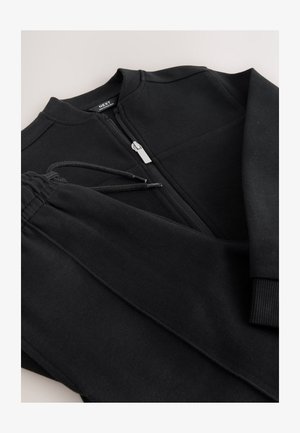 Survêtement sportif noir comprenant une veste zippée et un pantalon à cordon de serrage, fabriqué à partir d'un tissu texturé avec des poignets et une ceinture côtelés.