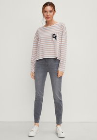 Maglietta a righe grigia e arancione a maniche lunghe con taschino sul petto, indossata con jeans skinny grigi e sneaker bianche. Vestibilità casual.