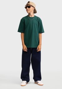 Grøn bomuld T-shirt med korte ærmer, kombineret med mørkeblå vidde-bukser og hvide sneakers med sorte striber. Beige kasket og solbriller.