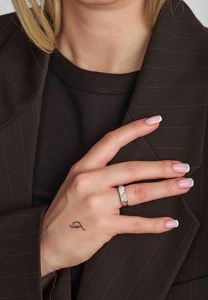 Hõbedane tekstuuriga sõrmus käel, millel on heleroosad küüned, toetudes pruuni triibulise jopetüki peale, all must t-särk ja nähtav tätoveering.