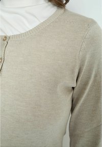 Cardigan beige realizzato in morbido tessuto a maglia, con scollatura tonda, chiusure con bottoni e dettagli di texture delicati lungo le cuciture.
