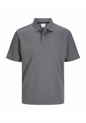 Mężczyzna w szarej koszulce polo z krótkim rękawem, czarnych podwiniętych dżinsach i czarnych wypolerowanych eleganckich butach, stoi na tle jednolitego jasnego tła.