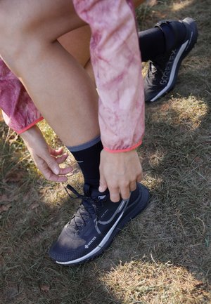 Osoba w różowej kurtce, wiążąca czarne buty do biegania Gore-Tex, kucając na suchej trawie na zewnątrz.