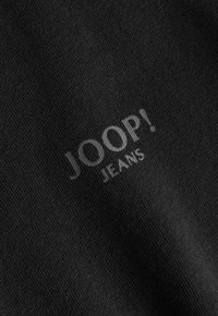 Tela negra con un logo sutil que dice "JOOP! JEANS", impreso en un tono más claro. La textura parece suave y de trama apretada.