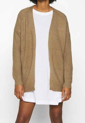 Persona che indossa un cardigan lavorato a maglia beige largo sopra un vestito bianco corto, in piedi contro uno sfondo semplice e chiaro.
