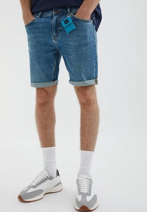 Homme portant un short en denim bleu retroussé avec un porte-clés turquoise, des chaussettes blanches et des baskets blanc-gris, debout les mains dans les poches.