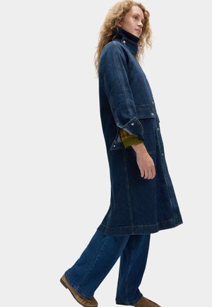 Dunkelblauer Denim-Trenchcoat mit hohem Kragen, Knopfverschlüssen, seitlichen Taschen und weiten Ärmeln, über einem grünen Pullover getragen.