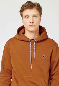 Bruine hoodie met een voorkuipezak, trekkoorden in het blauw met rode accenten, en een klein blauw boompje-logo op de linkerkant van de borst.