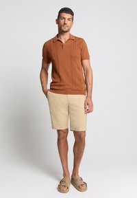 Polo shirt en maille marron avec un motif texturé, associé à un short beige clair. Modèle portant des sandales marron clair. Tenue simple et décontractée.