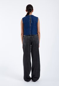 Chaleco de mezclilla recortado en azul oscuro con escote redondo y cremallera expuesta en la espalda, combinado con jeans de mezclilla negros de pierna ancha y cintura alta.