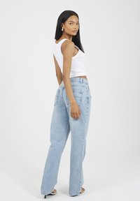 Kobieta z długimi czarnymi włosami ubrana w białą bezrękawnik, jasnoniebieskie spodnie z wysokim stanem i czarne sandały na obcasie, pozująca bokiem na białym tle.