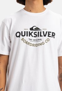 Camiseta blanca de algodón con un gran diseño gráfico en negro y dorado que presenta el texto "Quiksilver" y "Boardriding Co" en letras prominentes.