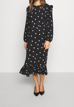 Czarna sukienka w białe groszki, z długimi bufkowymi rękawami i falbaną u dołu, wykonana z lekkiego materiału, zestawiona z czarnymi szpilkami o spiczastych noskach.