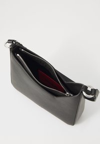 Sac en cuir noir avec fermeture éclair, de forme angulaire et accessoires en métal argenté. L'intérieur présente un porte-carte rouge avec un texte en relief.