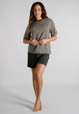 Frau steht barfuß, trägt ein kurzärmliges Oberteil mit Leopardenmuster und schwarze Shorts vor einem einfarbigen Hintergrund.