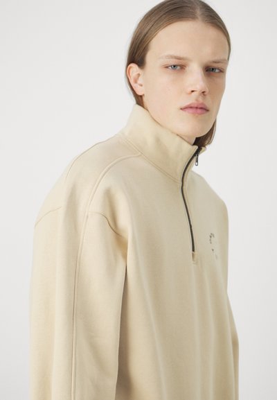 Felpa beige con zip a un quarto in tessuto morbido, caratterizzata da un collo alto e un piccolo dettaglio grafico sul petto. Vestibilità rilassata con dettagli delle cuciture.
