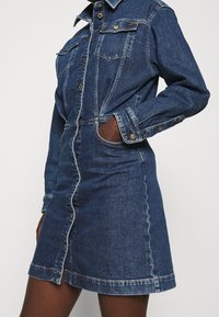 Jeanskleid mit tailliertem Oberteil und ausgestelltem Rock. Verfügt über eine Knopfleiste, zwei Brusttaschen und einen Kragen. Mittelblaue Waschung mit sichtbaren Nähten.