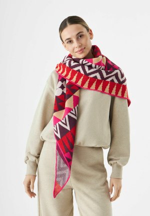 Vrouw in een beige casual outfit met een kleurrijke geometrische sjaal om haar nek, tegen een witte achtergrond.